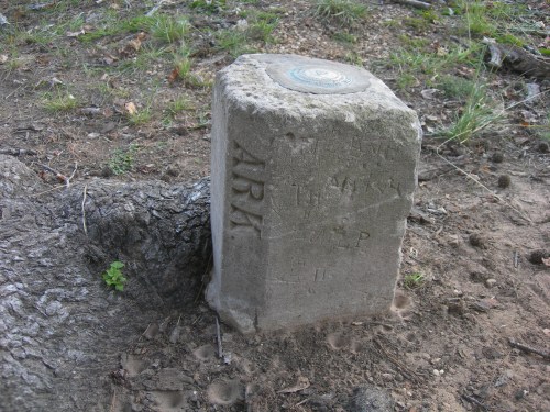 A weathered stone marker with engraved letters and a circular emblem, surrounded by dirt and grass.