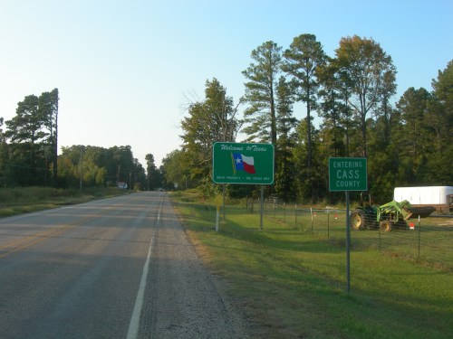 Welcome to Texas sign alongside a rural road, with a Cass County sign and greenery in the background.