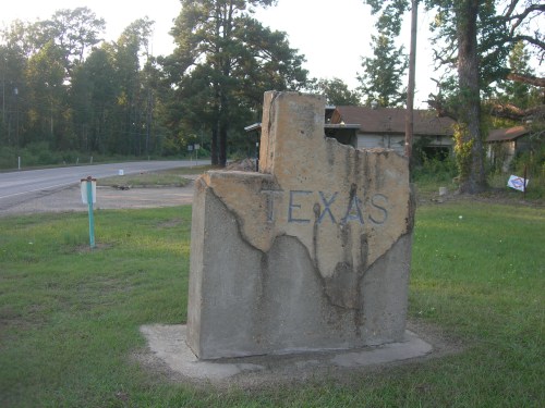 A concrete Texas-shaped monument with the word "TEXAS" engraved, set in a grassy area near a road.