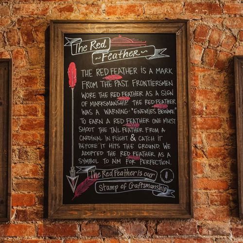 Chalkboard sign on a brick wall explaining the significance of the red feather in craftsmanship and marksmanship.