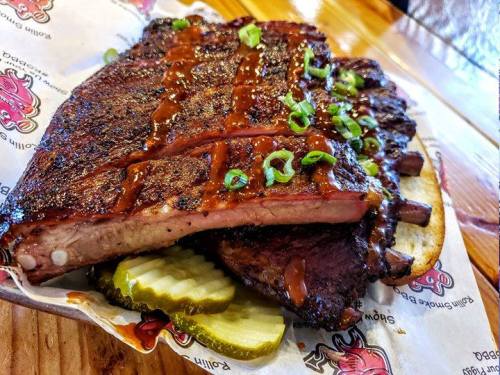 A plate of smoked beef ribs topped with barbecue sauce and green onions, served with pickles on the side.