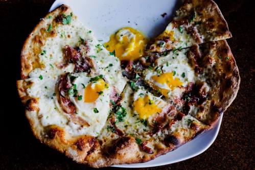 A pizza topped with eggs, herbs, and crispy meat, served on a white plate.