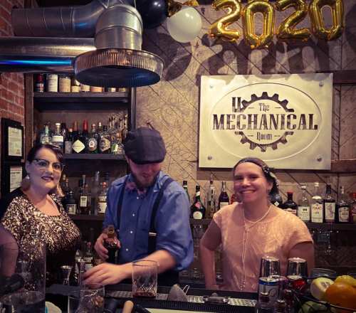Three bartenders at a bar, with a sign reading "The Mechanical Room" and "2020" balloons in the background.