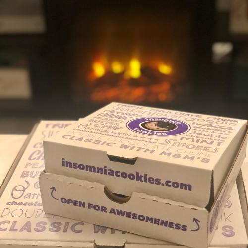 Two Insomnia Cookies boxes stacked in front of a cozy fireplace, featuring the text "Open for Awesomeness."