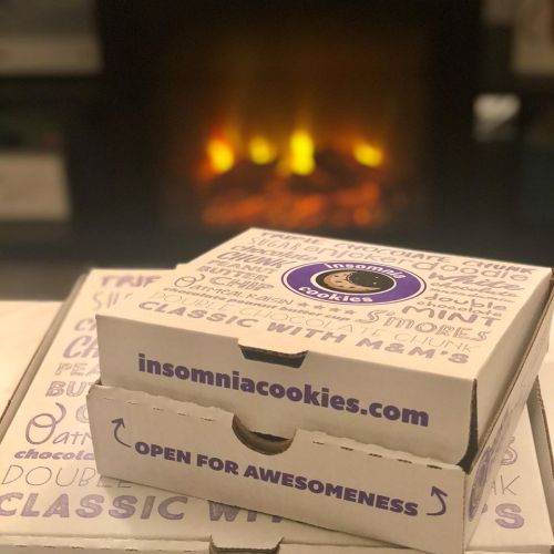 Two stacked cookie boxes from Insomnia Cookies in front of a cozy fireplace.