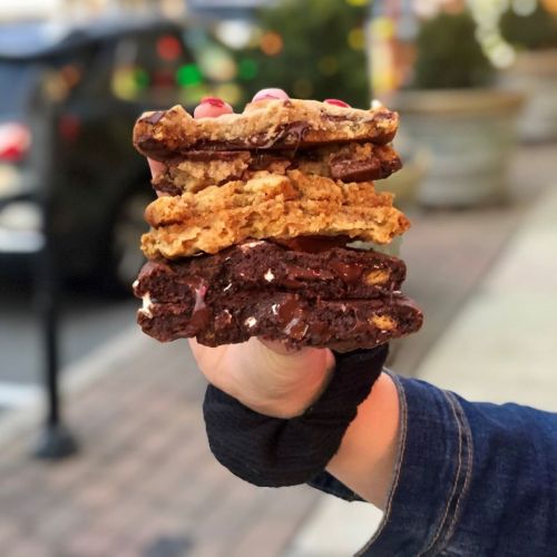 A hand holding a stack of three cookies: a chocolate chip, an oatmeal, and a brownie cookie, against a blurred background.