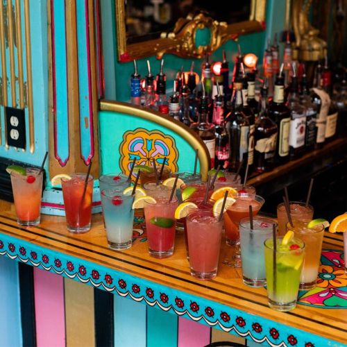 A colorful array of cocktails with garnishes on a vibrant bar, surrounded by various bottles in the background.
