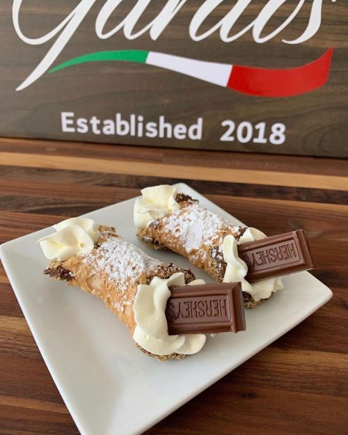 Two cannoli filled with cream, topped with whipped cream and a Hershey's chocolate bar, on a white plate.
