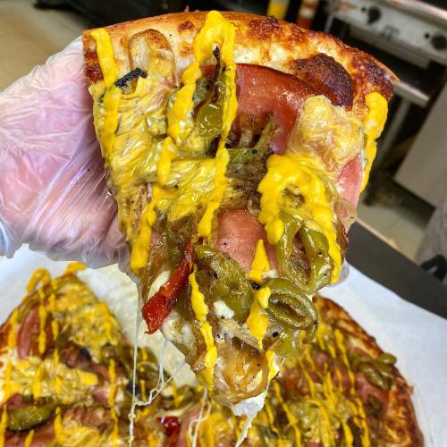 A hand holding a cheesy pizza slice topped with jalapeños, yellow mustard, and various toppings, with a whole pizza in the background.