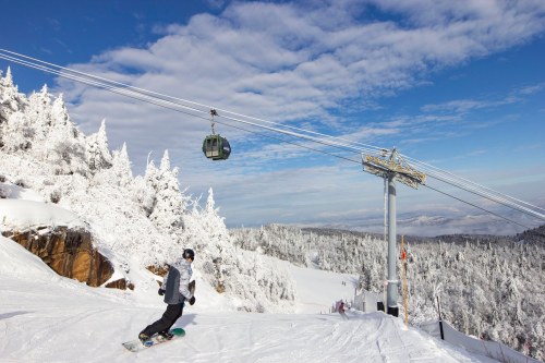 A snowboarder descends a snowy slope with a gondola lift in the background and a scenic mountain view.