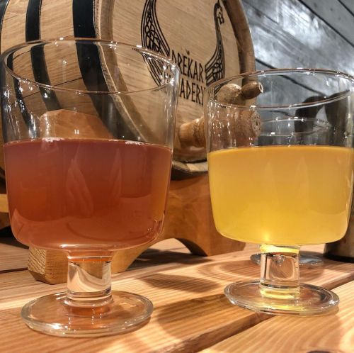 Two glasses of beverages, one pinkish-red and the other yellow, set on a wooden table near a barrel.