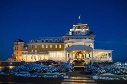 A grand yellow hotel with a festive wreath, illuminated at night, surrounded by snow and holiday decorations.