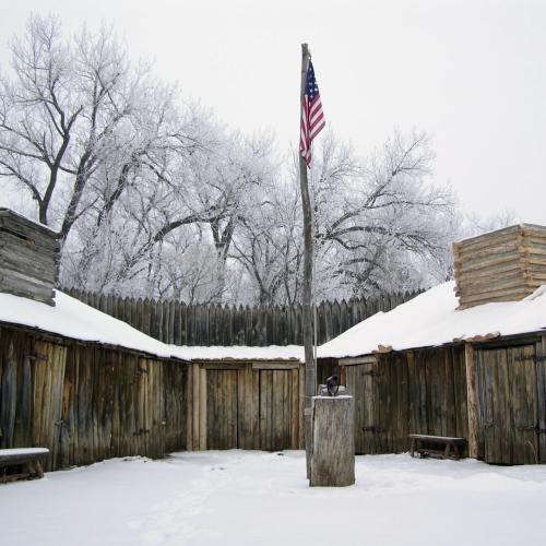 A snowy fort with wooden structures and an American flag flying in the center, surrounded by frosted trees.