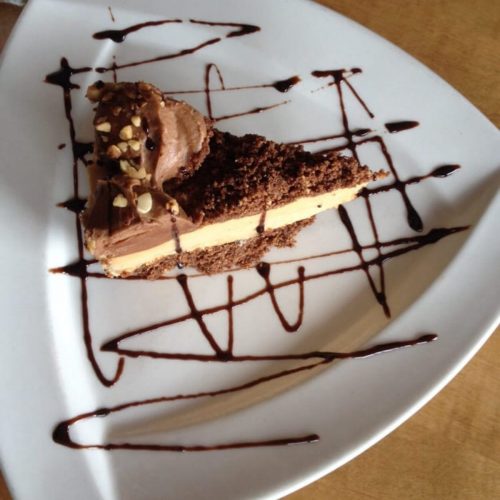 A slice of chocolate cake with creamy layers, topped with nuts, on a white plate drizzled with chocolate sauce.