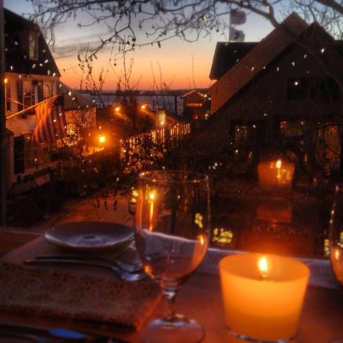 A cozy dining setting with a candle, glassware, and a sunset view over a charming street illuminated by lights.