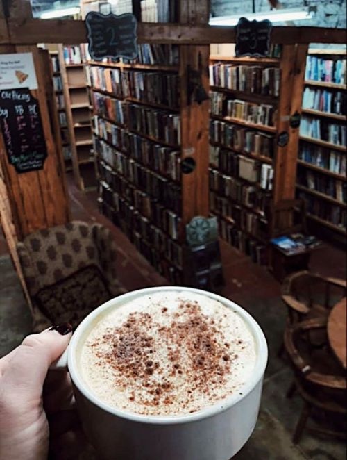 A hand holds a coffee cup topped with cinnamon, with a cozy library filled with books in the background.