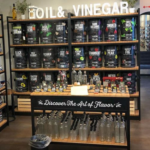 A display of various oils and vinegars on shelves, with a central table featuring glass bottles and a sign about flavor.