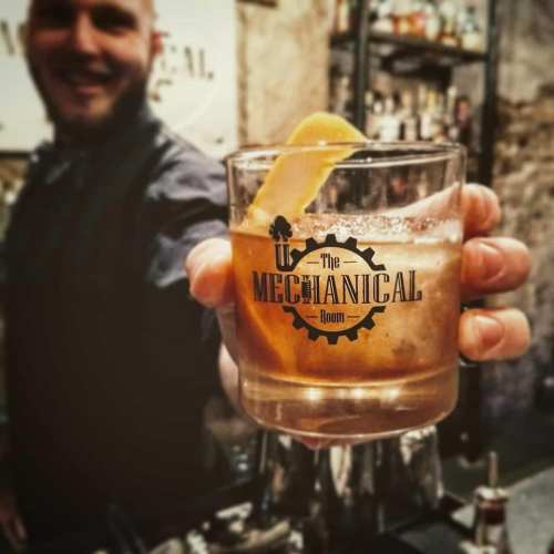 A bartender holds a glass with a cocktail, featuring a lemon slice, in a bar setting with shelves of drinks in the background.