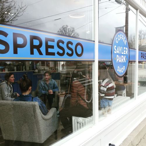A cozy coffee shop with people chatting inside, featuring a large window displaying the shop's name and logo.