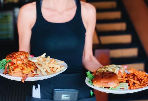 A server holds two plates of burgers with fries and sweet potato fries, standing in a restaurant.