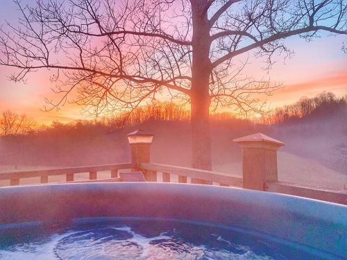 A serene sunset view from a hot tub, with mist rising and trees silhouetted against a colorful sky.