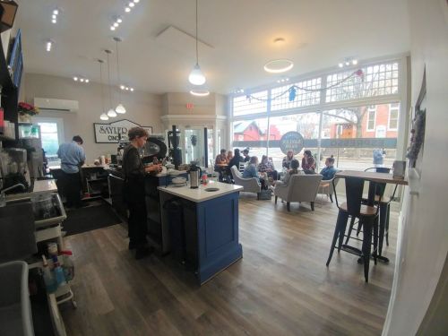 A cozy café interior with patrons seated, a barista at the counter, and large windows letting in natural light.