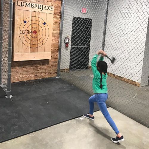 A child throws an axe at a wooden target in a fenced axe-throwing venue.