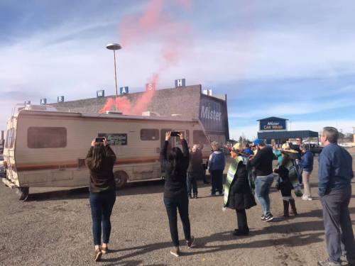 A crowd gathers around a vintage RV releasing pink smoke, with a car wash in the background.