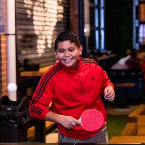A young boy in a red jacket smiles while playing table tennis indoors.
