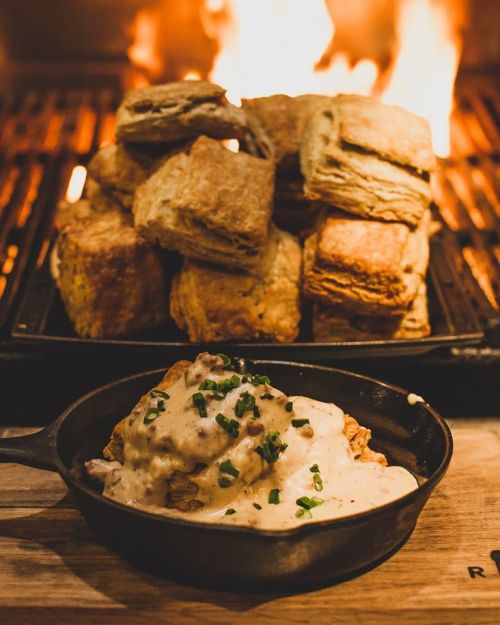 A cast iron skillet with creamy gravy and chopped chives, with a stack of golden biscuits and flames in the background.