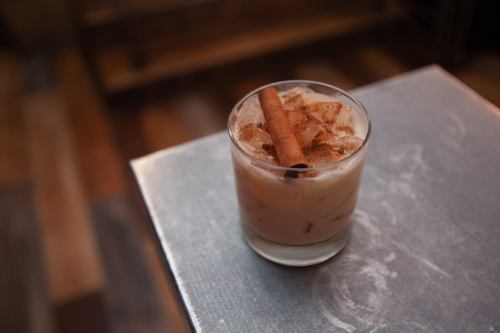 A glass of iced cocktail with a cinnamon stick on top, placed on a metallic surface.