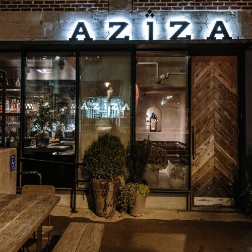 Exterior of Aziza restaurant at night, featuring a wooden door, large windows, and illuminated signage.
