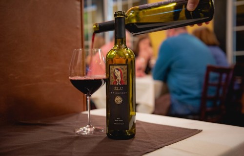 A bottle of red wine being poured into a glass at a restaurant table, with diners in the background.