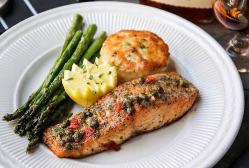 A plate featuring grilled salmon with capers, asparagus, a lemon wedge, and a baked potato.