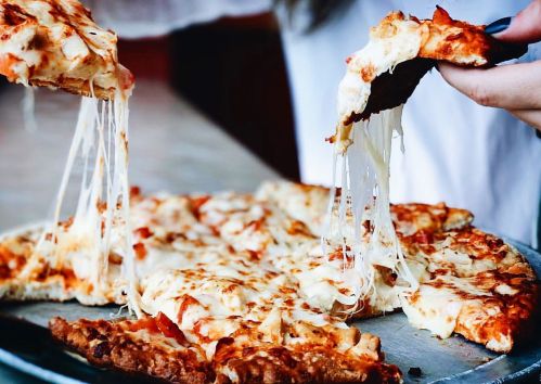 A close-up of a cheesy pizza being pulled apart, showcasing gooey cheese and toppings on a metal tray.