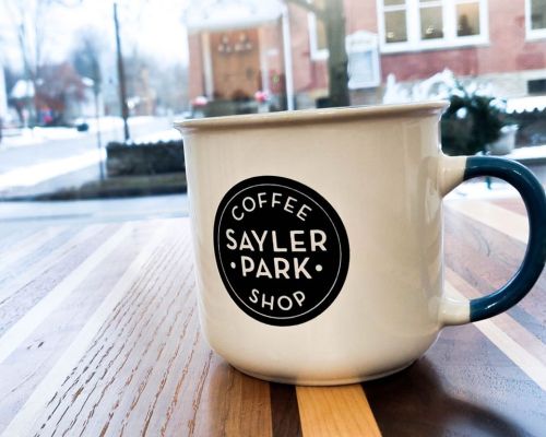 A coffee mug with "Sayler Park Shop" logo sits on a wooden table, with a snowy scene visible outside.