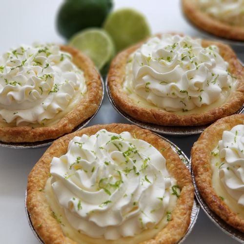 Four mini key lime pies topped with whipped cream and lime zest, with fresh limes in the background.