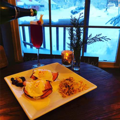 A cozy breakfast scene with eggs benedict, a cocktail, and a candle, set against a snowy window view.