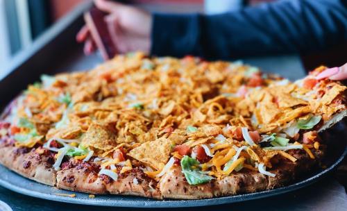 A large pizza topped with lettuce, tomatoes, cheese, and crunchy tortilla chips, with a hand reaching for a slice.