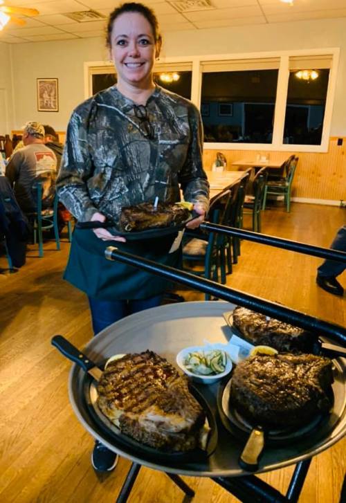 A server in camouflage holds two trays of large steaks, with a side of pickles, in a rustic dining setting.