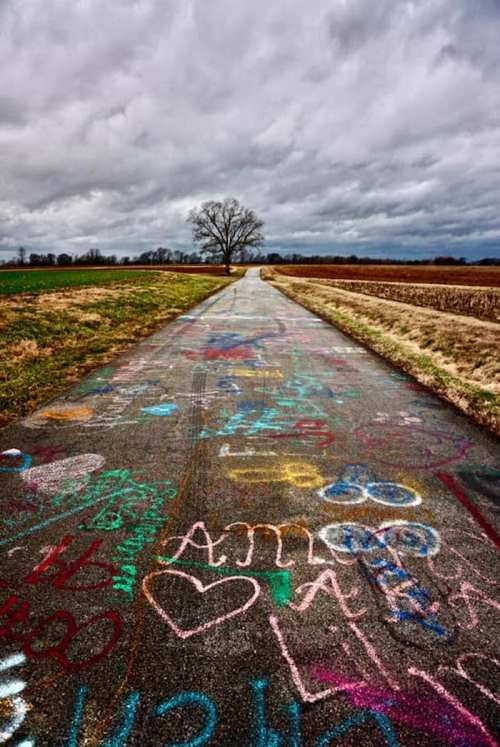 A colorful, chalk-covered road stretches into the distance, flanked by fields under a cloudy sky and a lone tree.