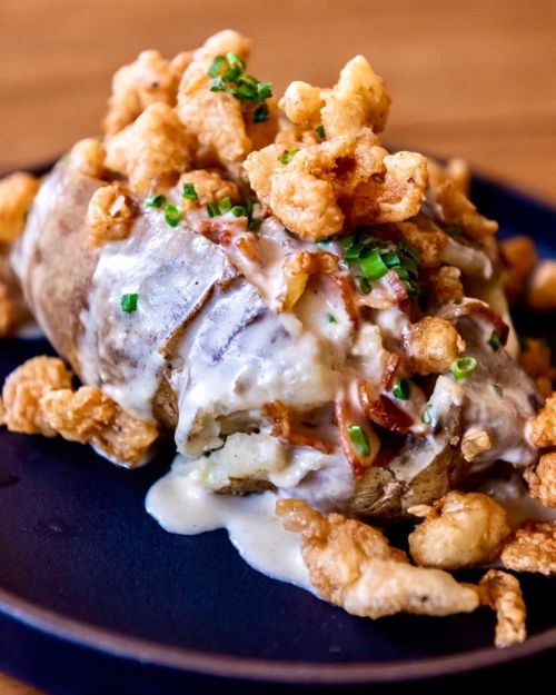 Baked potato topped with creamy sauce, chives, and crispy fried bits on a dark plate.