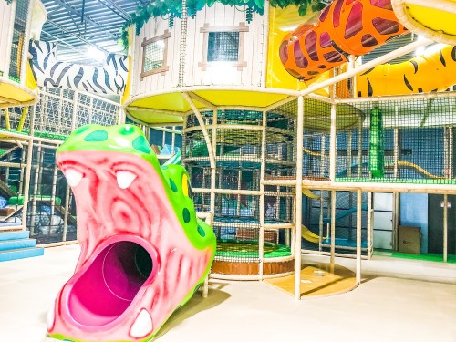Colorful indoor playground featuring a large green and pink slide shaped like a monster's mouth, with climbing structures.