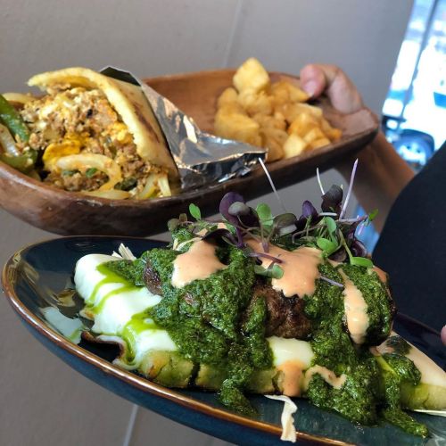 A plate with green enchiladas topped with sauce and microgreens, alongside a wooden dish of fried potatoes and a wrapped burrito.