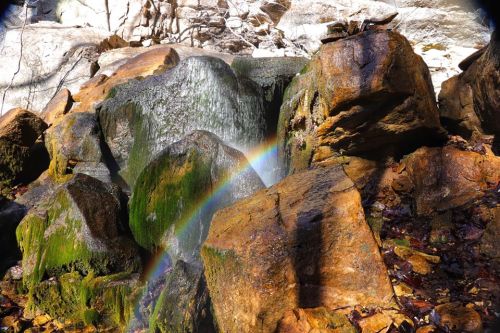 A vibrant rainbow arcs over moss-covered rocks near a waterfall, surrounded by natural stone formations.