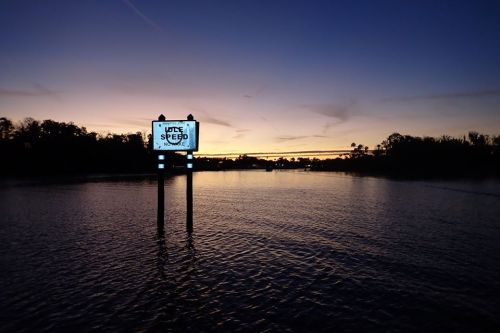 A sign displaying "SLOW SPEED" stands over calm water at sunset, with silhouettes of trees in the background.
