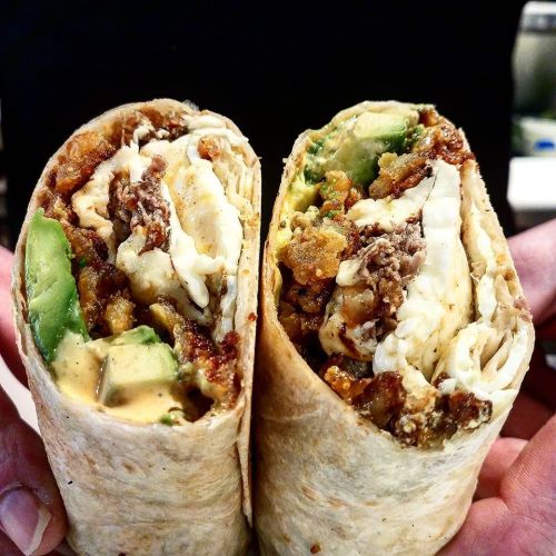 Two halves of a burrito filled with meat, avocado, and crispy ingredients, held by hands against a dark background.