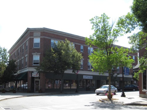A brick building on a corner with shops, trees, and a parked car under a clear sky.