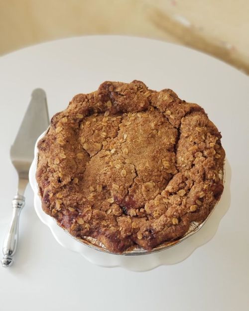 A freshly baked pie with a golden, crumbly topping sits on a white plate, accompanied by a silver pie server.