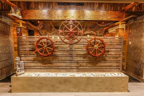 A rustic wooden wall featuring two ship wheels and decorative fishing nets, with a stone-filled planter at the base.